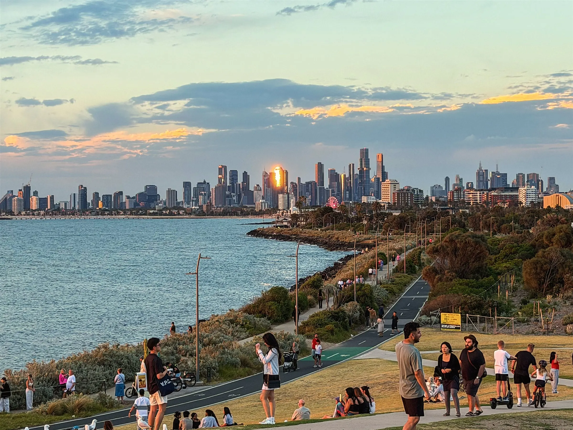 People gathered on a grassy foreshore at sunset, with the Melbourne skyline glowing across the bay
