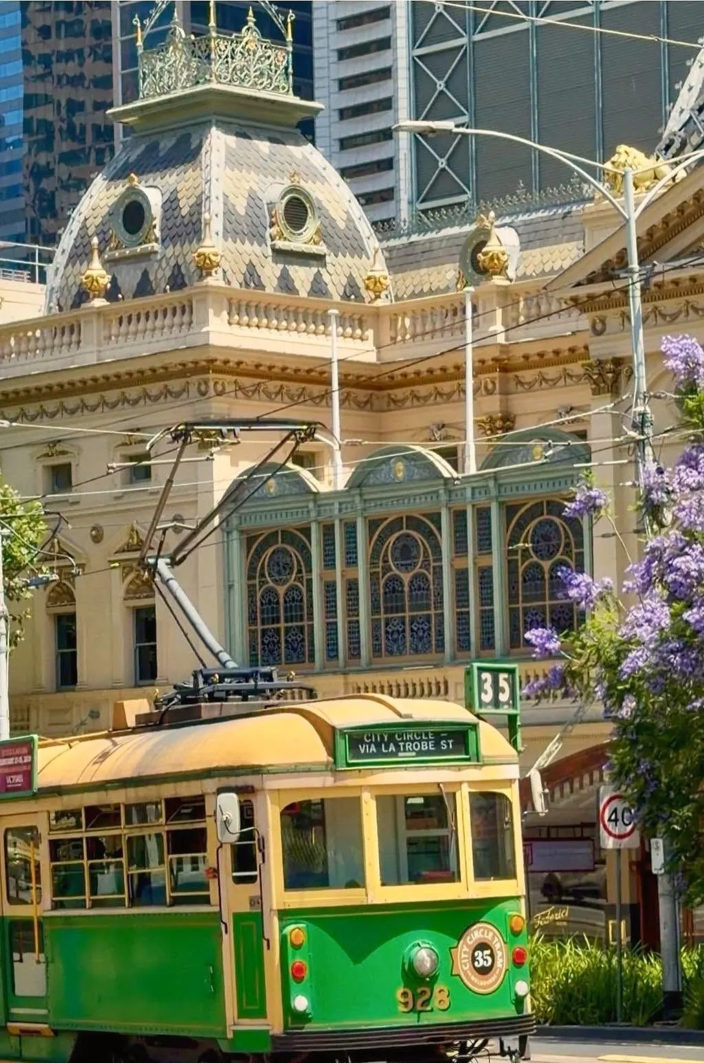 A green and cream City Circle tram passing the ornate domed Princess Theatre, with purple jacaranda flowers on the right