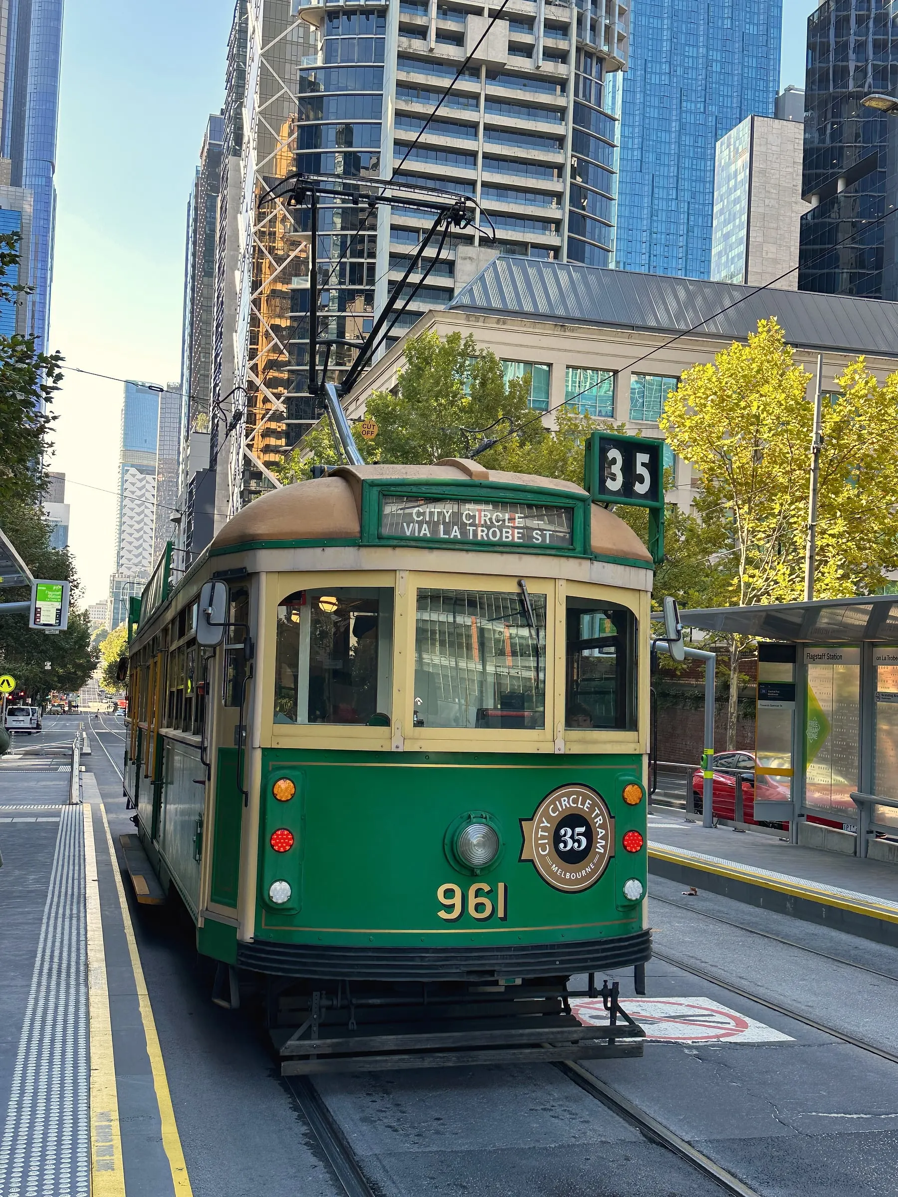 A green and cream heritage tram on the City Circle route, with city towers and a yellow autumn tree behind