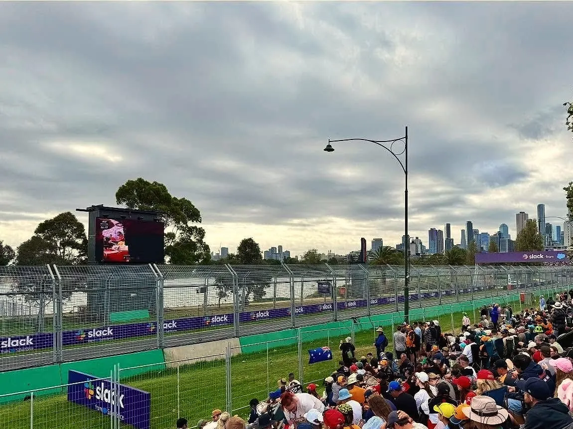 A big crowd seated on the grass beside the Grand Prix track at Albert Park, with the city skyline under grey clouds