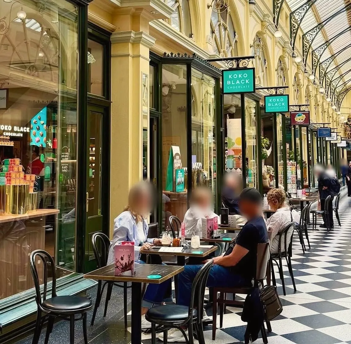People at café tables on the chequered floor of the Royal Arcade, beside shopfronts and green hanging signs