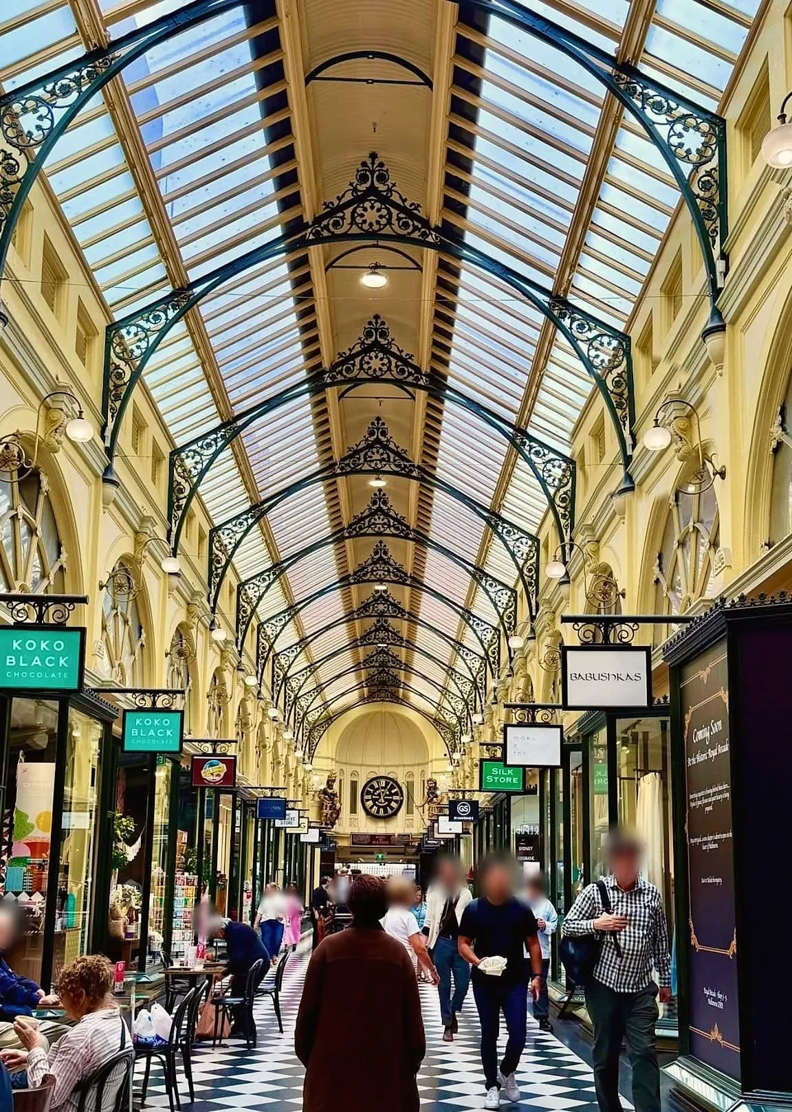 Looking down the Royal Arcade under its glass roof and iron arches, with a black and white tiled floor, shopfronts and people walking