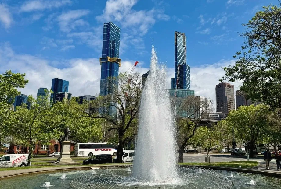 A tall fountain among leafy spring trees, with city towers and a blue sky behind