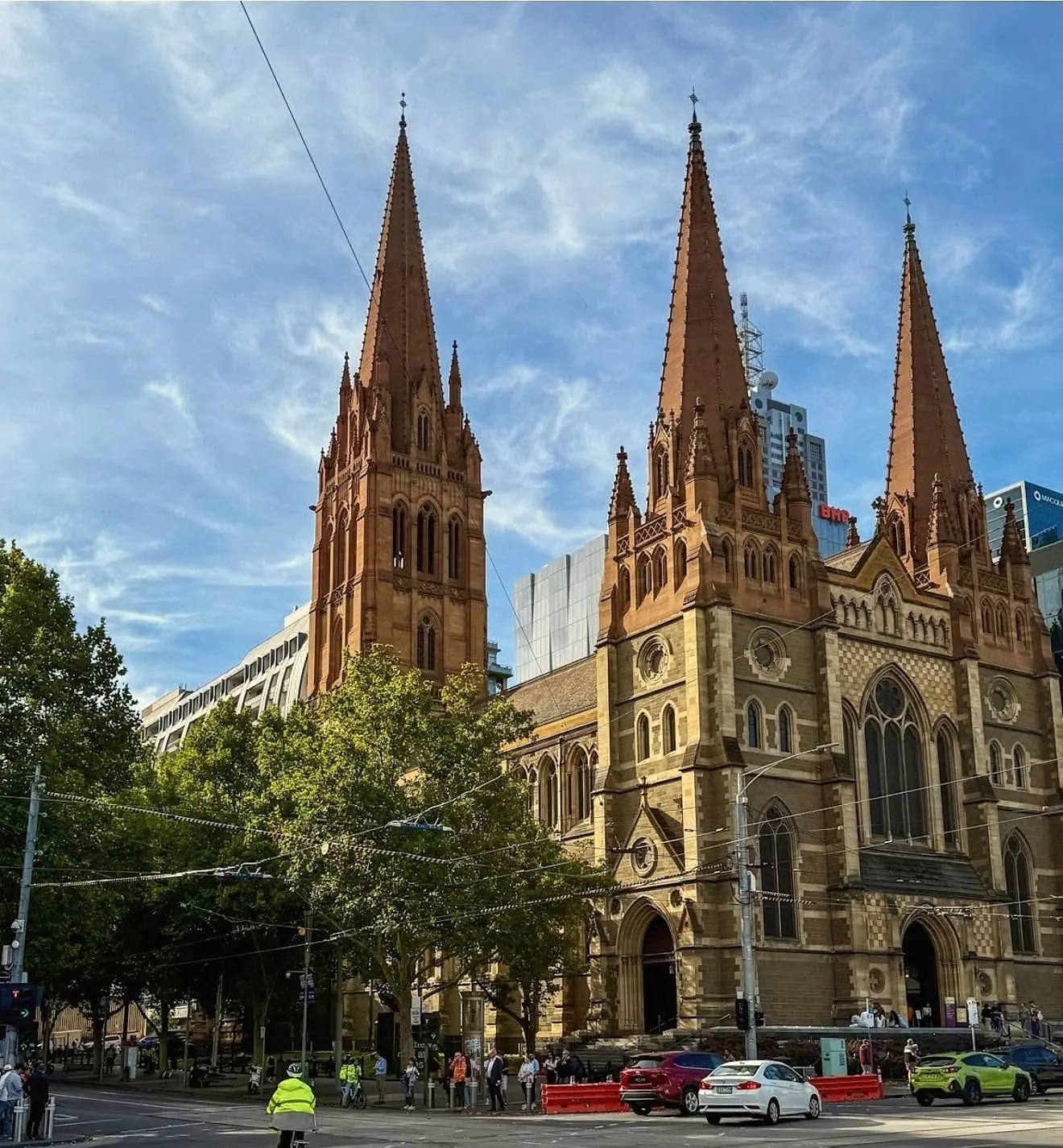 The three sandstone spires of St Paul’s Cathedral against a blue sky with wispy clouds, above street trees and a busy intersection