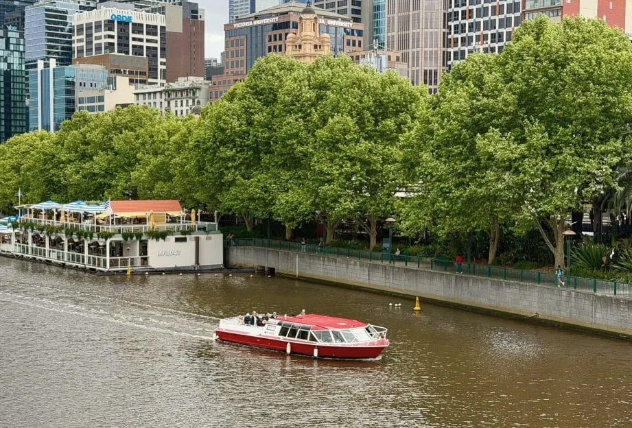 A red river cruise boat on the Yarra, beneath a row of leafy green trees with the city towers behind